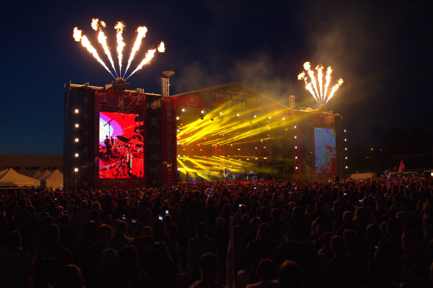 Eine große Menschenmenge bei einem Musikfestival schaut auf einer hell erleuchteten Bühne Performern zu, mit Zelten links und Feuerwerk in einem dunklen Himmel dahinter.
