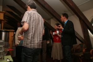 Mehrere Personen mit Tassen in der Hand, links ein Becher und ein Tisch, oben ein Dach und rechts Stühle.