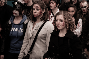 Eine Gruppe von Menschen in Zombie-Kostümen, einige in Kostümen, einer mit Hut und Handtasche, gehen eine Straße entlang mit einem Gebäude im Hintergrund.