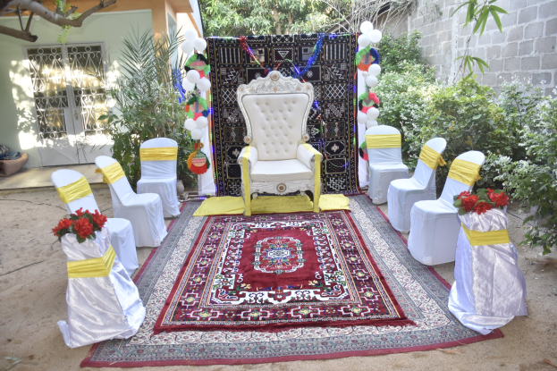 Eine Hochzeitszeremonie-Aufstellung in einem Garten mit weißen Stühlen, einem roten Teppich, Blumenbouquets, Dekorationsgegenständen, Luftballons, Pflanzen, Bäumen, einem Gebäude mit Fenstern und einer Tür und einer Wand auf der rechten Seite.