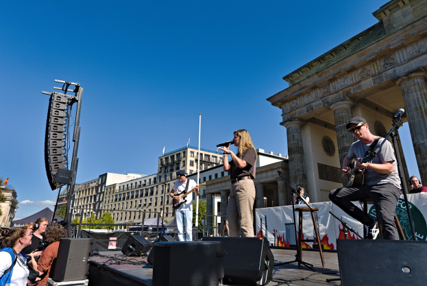 Eine Gruppe von Menschen, die auf einer Bühne vor dem Brandenburger Tor in Berlin Musik machen, umgeben von Lautsprechern und Equipment, vor einem klaren Himmel mit Gebäuden und Bäumen im Hintergrund.