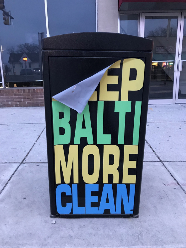 Schwarzer Müllcontainer mit dem Text "Keep Baltimore More Clean" auf einem Bürgersteig vor einem Gebäude mit Glastüren.