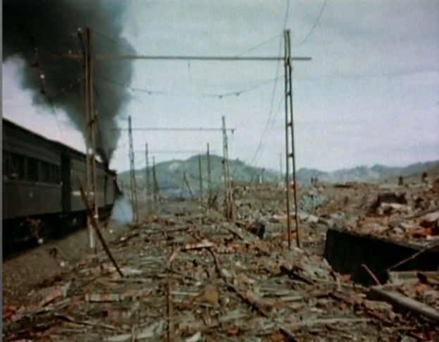 Ein Zug fährt durch eine verwöstete Landschaft mit Rauch, der aus ihm aufsteigt, umgeben von Strommasten und -leitungen, mit Höhen und einem klaren Himmel im Hintergrund und verstreuten Trömmern im Vordergrund.