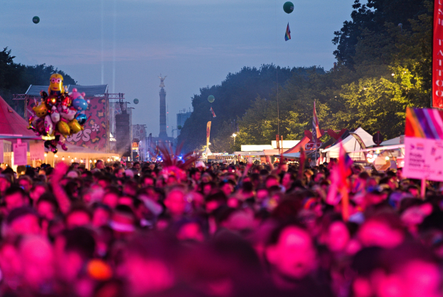 Große Menschenmenge auf einem nächtlichen Musikfestival mit farbigen Lichtern und Ballons, umgeben von Zelten, Bannern, Fahnen, Bäumen, einem Turm und einem sternenklaren Himmel.