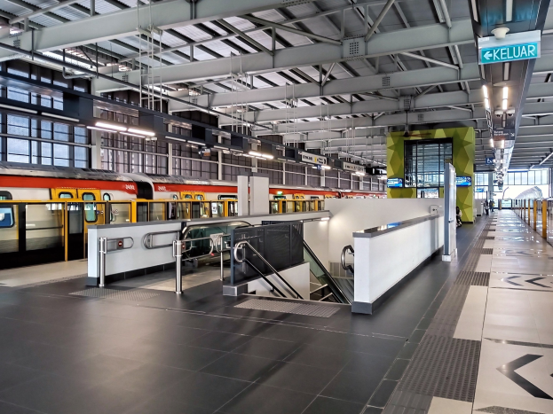 Ein Bahnsteig mit einem Zug auf den Schienen, eine Plattform mit Geländern, Treppen mit Handläufen, Informationsanzeigen, Deckenleuchten und Glaswände.