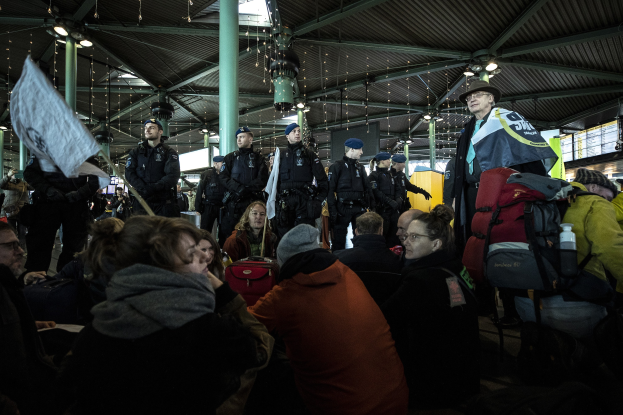 Eine Gruppe von Menschen steht vor einer größeren Menge in einem, was wie ein Bahnhof aussieht, mit Säulen und Deckenleuchten im Hintergrund; einige in der Menge tragen Mützen, tragen Taschen und halten Schilder, was auf eine Demonstration hindeutet.