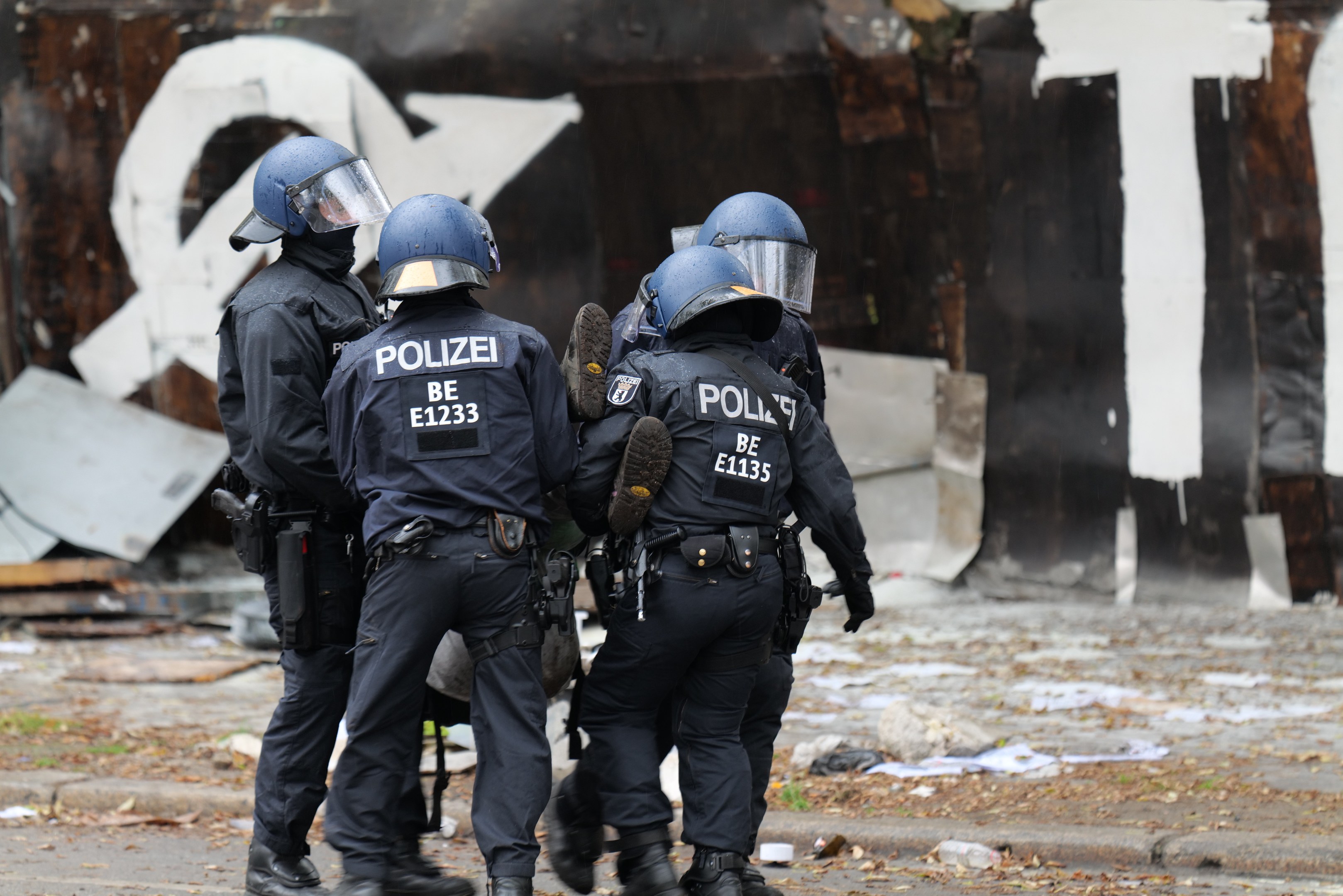 Polizeibeamte in schwarzen Uniformen und blauen Helmen stehen vor einem mit Graffiti bedeckten Gebäude während einer gewaltsamen Demonstration, mit verstreuten Papieren und Gegenständen auf dem Boden.