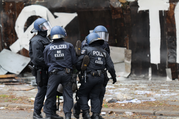 Polizeibeamte in schwarzen Uniformen und blauen Helmen stehen vor einem mit Graffiti bedeckten Gebäude während einer gewaltsamen Demonstration, mit verstreuten Papieren und Gegenständen auf dem Boden.