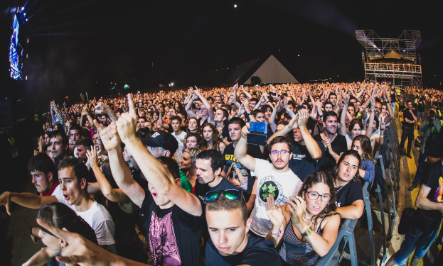 Eine große Menschenmenge auf einem Musikfestival mit erhobenen Händen, sitzend und stehend, hinter einem Metallzaun, mit Gebäuden und Lichtern im Hintergrund unter einem dunklen Himmel.
