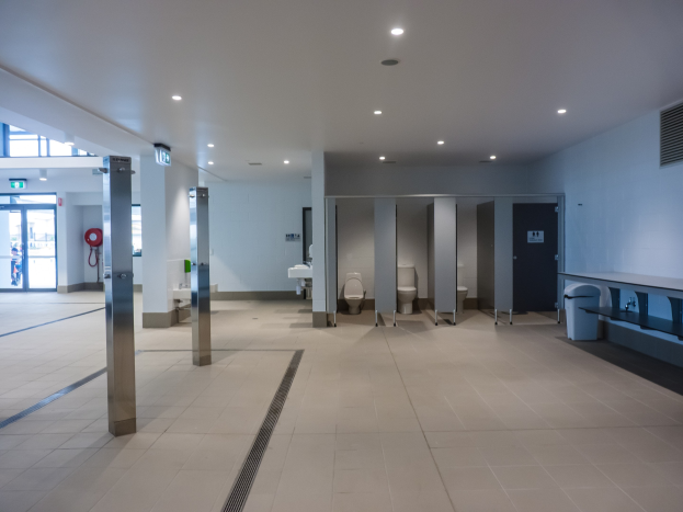 Große öffentliche Toilette mit mehreren Toiletten, tragenden Säulen, einem Mülleimer, einer Tür, einem Schild, einem Feuerlöscher und deckengehängten Leuchten.