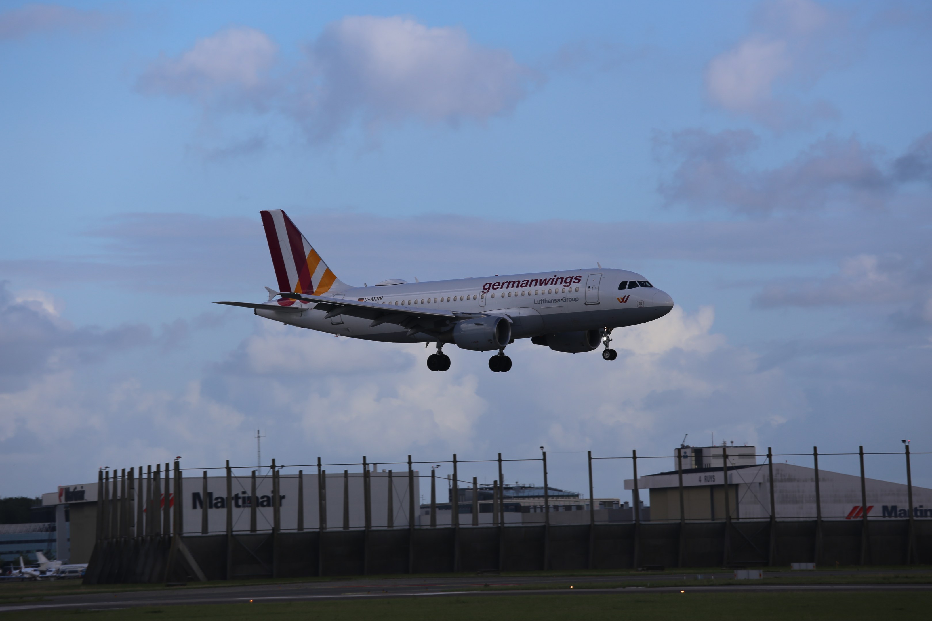 Germanwings Airbus A320-200 beim Start von Frankfurt Airport mit Gras darunter, einem Zaun im Vordergrund, Gebäuden und Bäumen im Hintergrund und einer bewölkten Himmel.