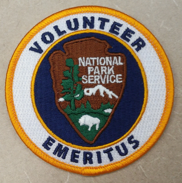 Volunteer National Park Service Emeritus Abzeichen mit weißem Zentrum, blauem Rand und gelbem Innenkreis.
