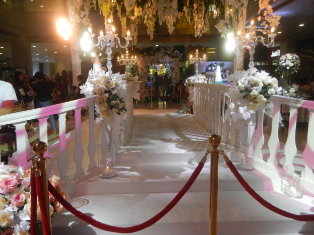 Eine Trauung in einem Einkaufszentrum mit weißem Teppich, Geländern, Blumenschmuck und Deckenbeleuchtung, bei der zahlreiche Gäste anwesend sind.