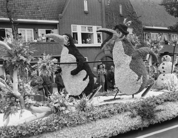 Schwarz-weiß-Foto eines Paradewagens mit Blumen und Pinguin-Figuren, umgeben von Zuschauern, Bäumen und Gebäuden.