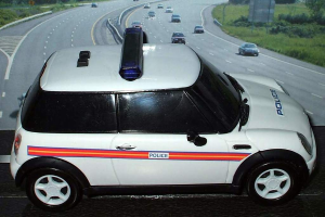 Ein weißer Polizeiwagen steht im Vordergrund mit mehreren fahrenden Fahrzeugen auf der Straße dahinter, umgeben von Gras und Bäumen in einer ländlichen Umgebung.