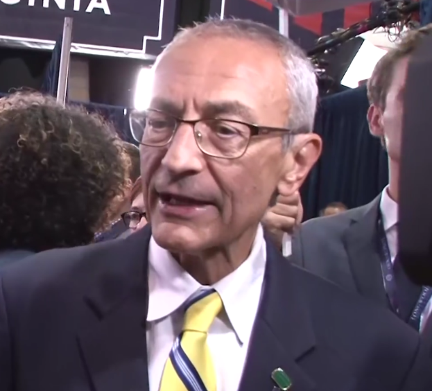 Ein Mann im Anzug und Krawatte, der eine Brille trägt, spricht mit Reportern vor einem Mikrofonständer, umgeben von einer Gruppe von Menschen, mit Scheinwerfern und einer Tafel im Hintergrund.