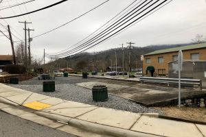 Eine Straßenszene in der Stadt mit Gebäuden, Fahrzeugen, Bäumen, Wolkenhimmel, einem Gehweg, Mülltonnen, einer Straßenlaterne, Strommasten mit Drähten.