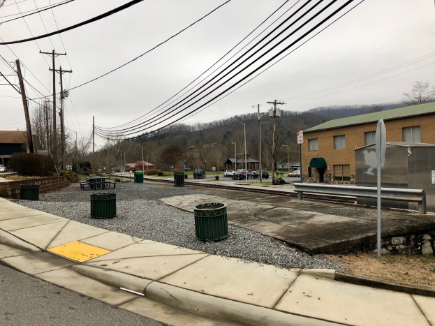 Eine Straßenszene in der Stadt mit Gebäuden, Fahrzeugen, Bäumen, Wolkenhimmel, einem Gehweg, Mülltonnen, einer Straßenlaterne, Strommasten mit Drähten.