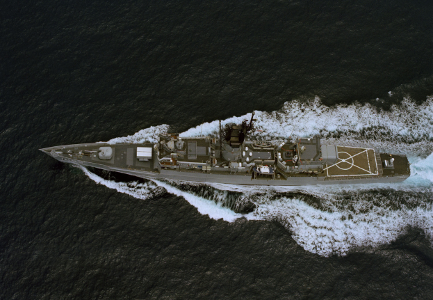 Luftaufnahme einer lenkwaffenbestückten Fregatte auf dem Ozean mit glitzerndem Sonnenlicht auf dem Rumpf und Wellen, die gegen den Schiffsrumpf schwappen, mit einem langen Rumpf, einem großen Geschützturm und blähenden Segeln.