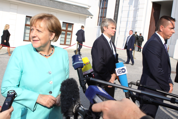Bundeskanzlerin Angela Merkel spricht vor dem Kanzleramt in Berlin mit Reportern, umgeben von Menschen mit Mikrofonen, mit Gebäuden im Hintergrund.