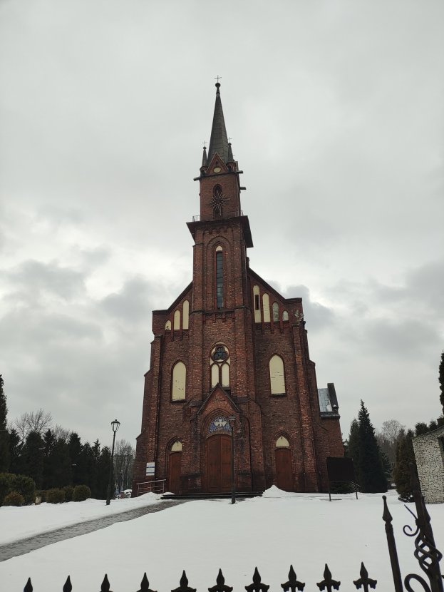 Kirche mit einem Turm, der in einem verschneiten Feld steht, von einem Metallzaun umgeben, umgeben von Straßenmasten, Bäumen und Pflanzen unter einem bewölkten Himmel.
