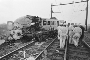 Ein Schwarz-Weiß-Bild einer Zugkollision mit Menschen in Helmen drumherum, Strommasten, Bäume, Gebäude und einen klaren Himmel im Hintergrund.