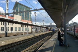 Eine Bahnhofsszene mit Personen, die rechts stehen und sitzen, einem Zug in der Mitte und Gebäuden, Pfählen und einem bewölkten Himmel im Hintergrund.