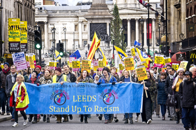 Gruppe von Menschen marschiert mit einem 'Stand Up to Racism'-Schild die Straße entlang, mit Gebäuden, Bäumen, Laternenmasten, Verkehrsampeln und einer Statue im Hintergrund bei bewölktem Himmel.