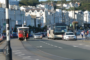Eine belebte Stadtstraße mit hohen Gebäuden, Fahrzeugen, Fußgängern, Ampeln, Laternen, Bäumen und einem klaren blauen Himmel.