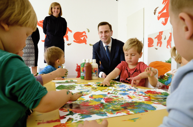 Kinder sitzen um einen Tisch mit Farbe an den Händen, Papieren und Flaschen, während eine lächelnde Frau im Hintergrund steht und Gemälde an der Wand hängen.