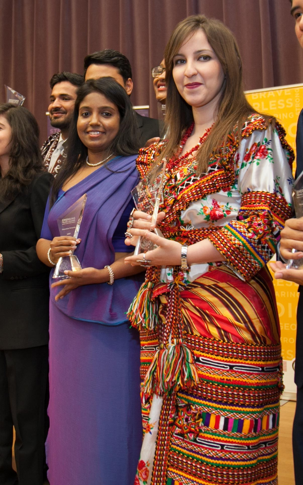 Eine Gruppe von Menschen auf einer Bühne mit einer Frau in der Mitte, die eine Trophäe hält, vor einem Vorhang und einer Tafel mit der Aufschrift "Bravo Segen Award Gewinner."
