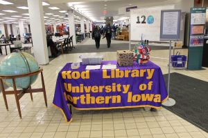 Eine Expo-Szene mit einem Tisch, auf dem verschiedene Landesflaggen, eine Box, Papiere und ein Banner liegen, eine Kugel links daneben, ein Anschlagbrett rechts daneben und mehrere Tische und Stühle im Hintergrund.
