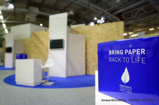 Ein Messestand mit einem blauen und weißen Schild, auf dem 'Bring Paper Back to Life' steht, auf der rechten Seite. Im Hintergrund sind Stühle, Tische, ein Fernseher und Deckenleuchten zu sehen, alles leicht unscharf.