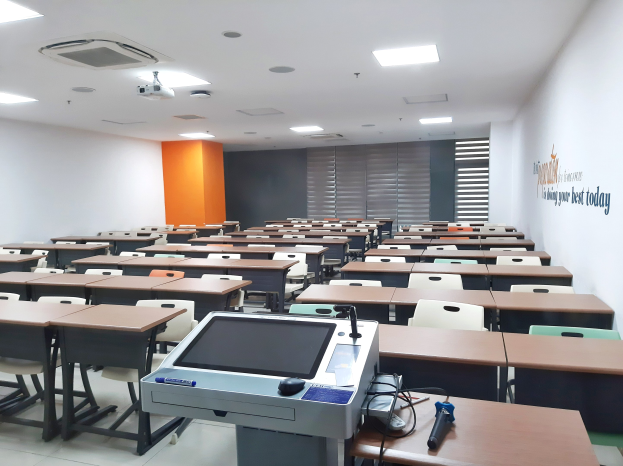 Ein Klassenzimmer mit Tischen und Stühlen in Reihen, ein deckengehängter Projektor und Gegenstände auf Tischen, mit Text an der rechten Wand, der auf eine Privatschule in Singapur hinweist.