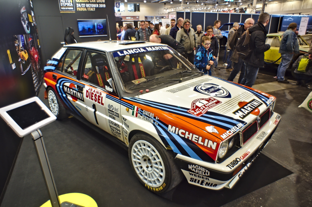 Ein Lancia Delta HF Integrale Evoluzione Rallyefahrzeug auf einer Autauschstellung umgeben von Menschen in bunten Kleidern, mit einem Bildschirmständer links und Deckenleuchten und Tafeln im Hintergrund.