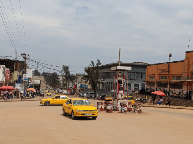 Ein gelbes Taxi fährt eine belebte Stadtstraße entlang, gesäumt von Gebäuden, während Fußgänger mit Regenschirmen auf dem Gehweg darunter unter bewölktem Himmel gehen, mit Strommasten und Drähten im Hintergrund.
