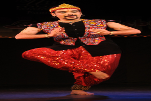 Ein Mann in roter und schwarzer Kleidung tanzt auf einer Bühne mit dunklem Hintergrund, sein Gesicht ist sichtbar mit geschlossenen Augen und leicht geöffnetem Mund.