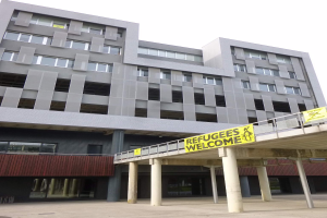 Ein großes Gebäude mit einem Schild, auf dem "Flüchtlinge Willkommen" steht, umgeben von Säulen und einem Geländer, mit dem Himmel im Hintergrund.