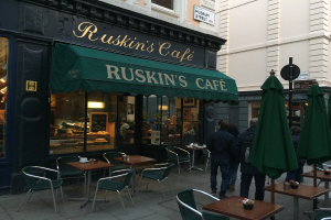 Eine Gruppe von Menschen steht vor einem Café mit der Aufschrift 'Ruskin's Cafe'.