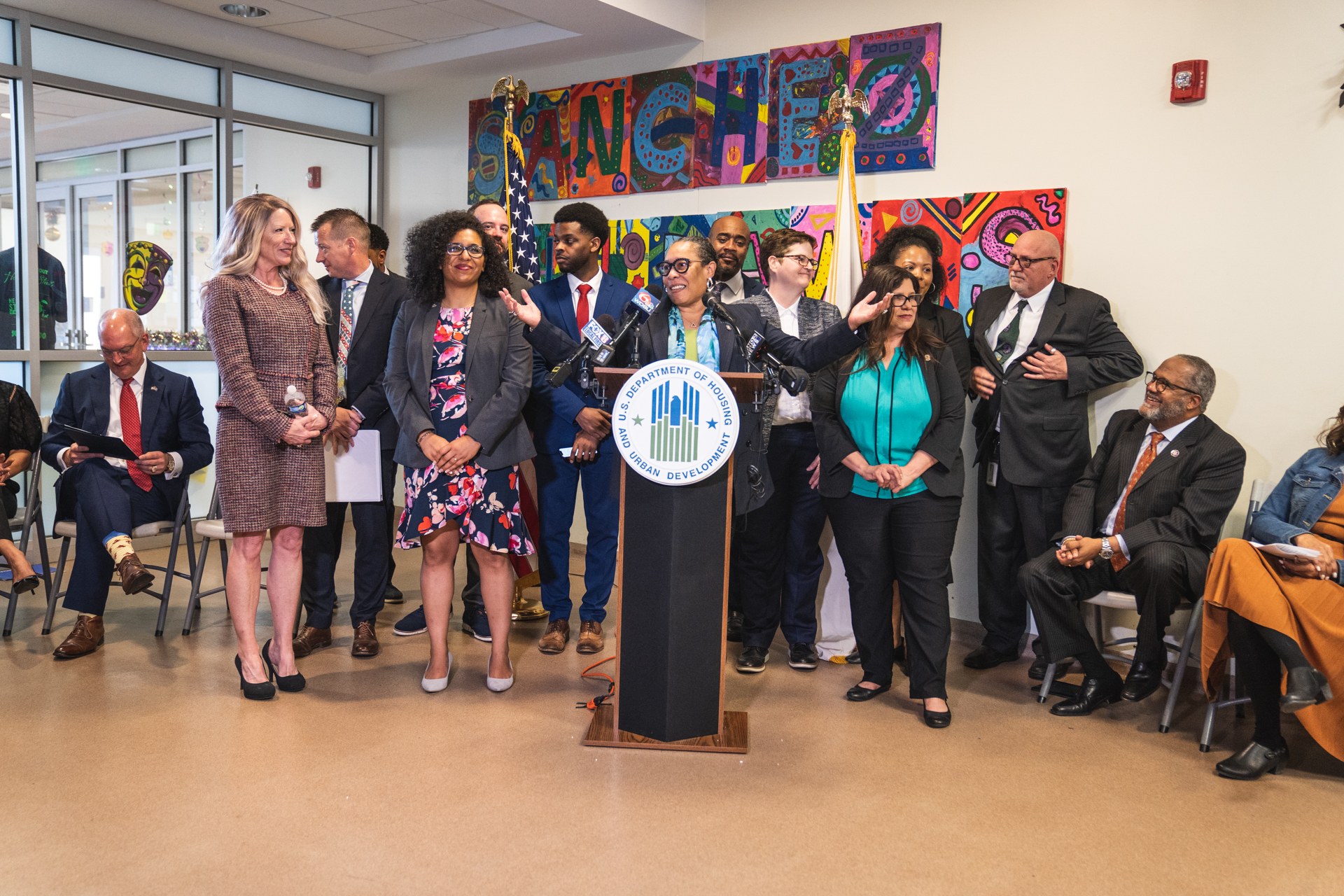 Gruppe von Menschen in einem Raum mit einem Rednerpult, Mikrofon und einer Tafel mit der Aufschrift "Sancho-Stadtrat tritt dem Stadtrat bei", umgeben von Flaggen, Feuerlöscher und Deckenleuchten.