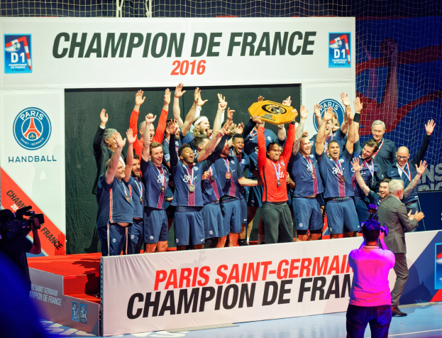 Eine Gruppe von Männern auf einer Bühne, die einen Pokal halten, mit einer Person, die filmt, und einem 'Champion de France 2016'-Schild im Hintergrund.