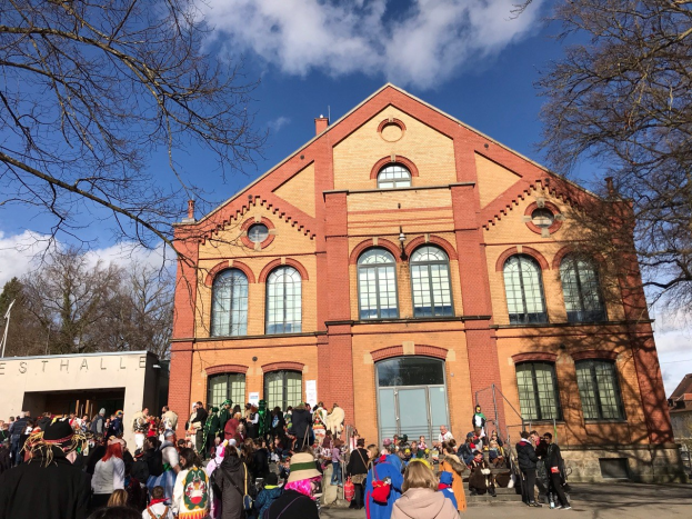 Eine große Gruppe von Menschen steht vor einem mehrstöckigen Gebäude mit Fenstern und Türen, umgeben von Bäumen unter einem bewölkten Himmel, einige tragen Mützen und Taschen.
