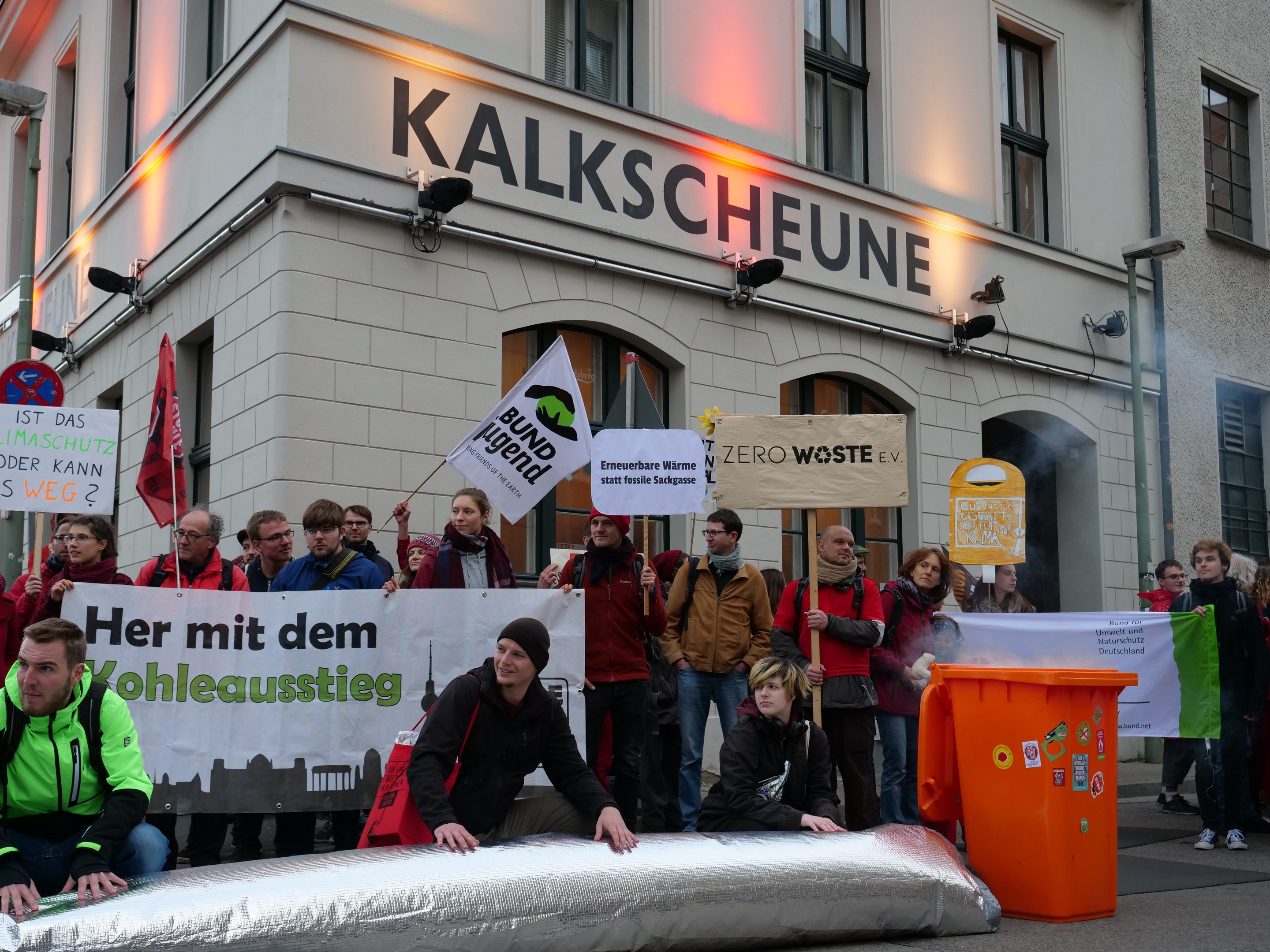 Eine Gruppe von Menschen steht vor einem Gebäude, hält Schilder und Plakate in der Hand, mit zwei Personen im Vordergrund und einem Müllcontainer auf der rechten Seite, während einer Protestaktion in Deutschland.