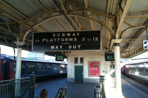 Zwei Züge stehen neben einem Bahnsteig mit zwei Bildschirmen und mehreren Schildern, die von der Decke hängen, eine Person steht in der Nähe einer Treppe und eine Tür ist an der Wand zu sehen.