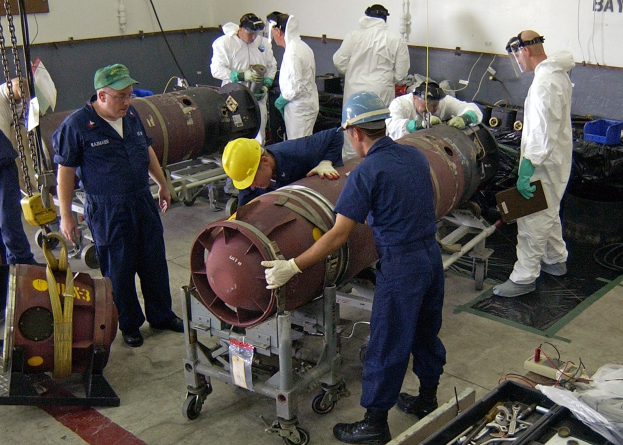 Männer in Helmen und Handschuhen zerlegen eine große Bombe in einer industriellen Fabrikumgebung mit Maschinen und Wandtext.