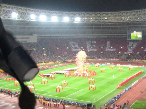 Ein Stadion mit Flutlicht, Eisengittern, Zuschauern, stehenden Personen, Werbetafeln, Anzeigetafeln, überwachungskamera, Fahnen und Deko.