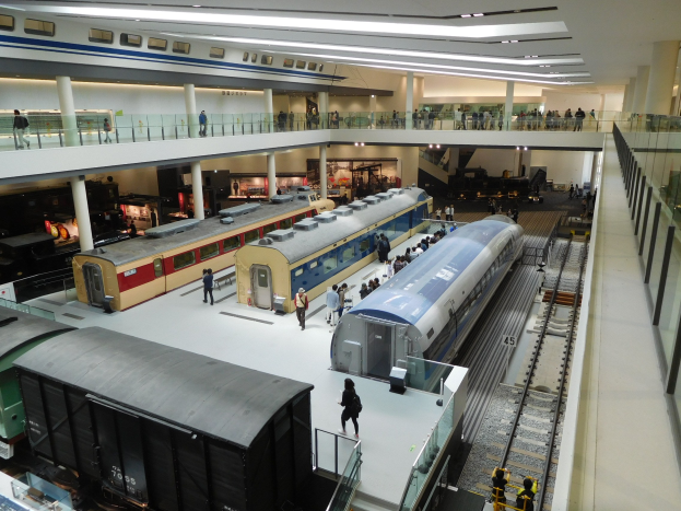 Eine große Museumsausstellung zeigt verschiedene Züge in unterschiedlichen Größen und Farben, mit Menschen, die vom Boden, Geländern und Säulen aus beobachten, unter Deckenlampen.