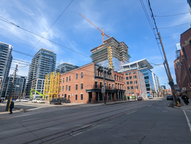 Eine belebte Stadtstraße in Toronto mit fahrenden Fahrzeugen, Fußgängern auf dem Gehweg, Strommasten mit Drähten, Schildern, Gebäuden und einer Baustelle mit einem Kran im bewölkten Himmel.