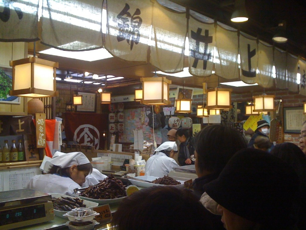 Restaurantmitarbeiter bereiten Gegenstände hinter einer Theke vor, während Kunden davor stehen, mit Flaschen, Menüs und Kleidung im Hintergrund.