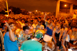 Menschen tanzen vorne bei einem Konzert mit Lichtern, Pfählen, Bäumen und anderen Gegenständen im Hintergrund.